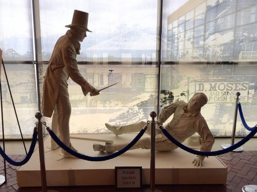 A white statue of a man in a top hat pointing a cane at another man lying on the ground, surrounded by ropes.