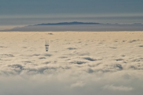 A tall tower rises above a sea of clouds, with a distant mountain range visible in the background.