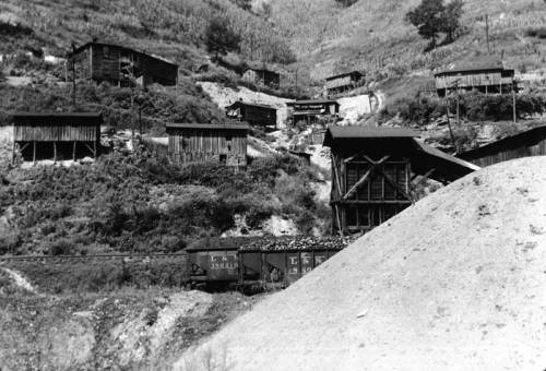 Black and white image of wooden mining structures on a hillside, with a train car and a large pile of earth in the foreground.