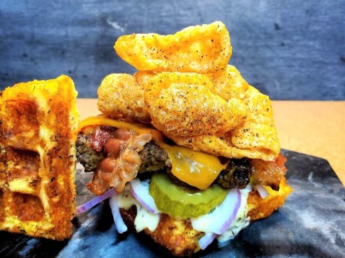 A stacked burger with bacon, cheese, pickles, and crispy pork rinds, served on a waffle bun.