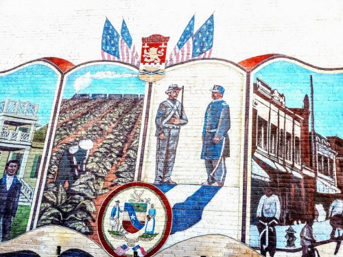 A colorful mural depicting historical scenes, including soldiers, farming, and local architecture, with flags and a coat of arms.
