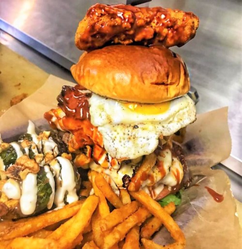 A towering burger with fried egg, spicy chicken, and fries on the side, served on parchment paper.