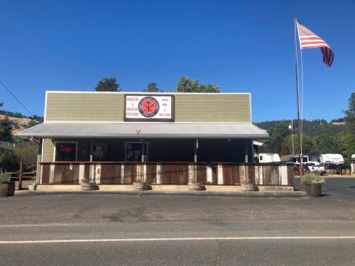 A small green building with a sign reading "Open," an American flag, and planters along the porch.