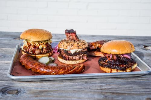 A tray of gourmet burgers and sausage, featuring various toppings and sides, on a rustic wooden table.