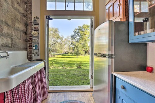 A bright kitchen with a vintage sink and blue cabinets, opening to a lush green outdoor view through a glass door.