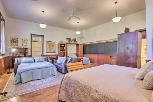 Spacious room with two beds, a wooden table, bookshelves, and a chalkboard, featuring warm lighting and wooden floors.