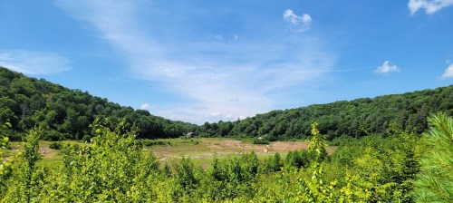 A panoramic view of a lush green valley surrounded by hills under a bright blue sky with scattered clouds.
