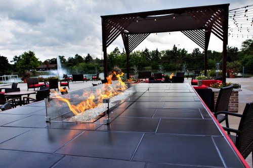 A modern outdoor patio with a fire feature, surrounded by chairs and greenery under a cloudy sky.