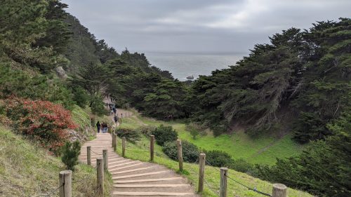 A winding path leads down through greenery towards the ocean, with people walking in the distance under a cloudy sky.