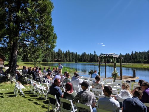 A scenic outdoor wedding setup by a lake, with guests seated on white chairs under a clear blue sky.