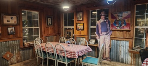 A rustic restaurant interior with wooden walls, checkered tablecloths, and a life-size cowboy cutout.