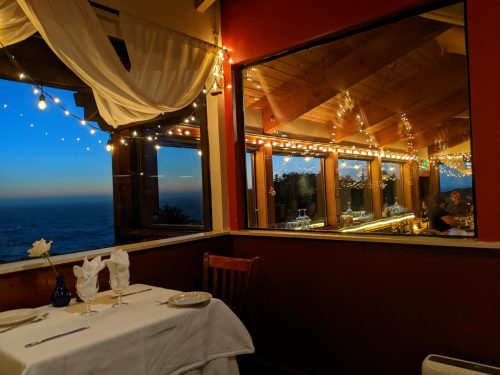 Cozy restaurant setting with a table for two, ocean view, and warm string lights illuminating the interior.