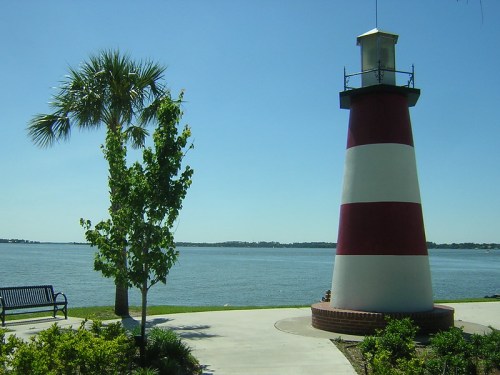 A red and white striped lighthouse beside a calm body of water, with palm trees and a bench in the foreground.