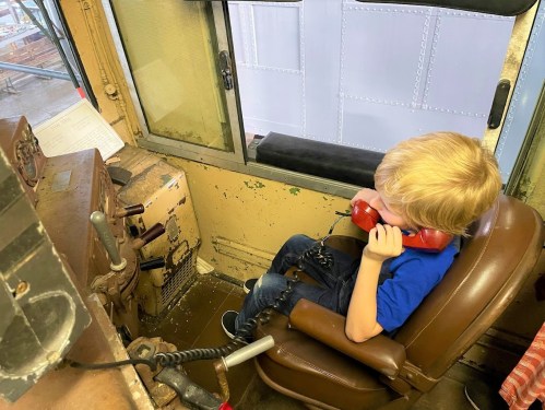 A young child sits in a train cab, holding a red phone, looking out the window.
