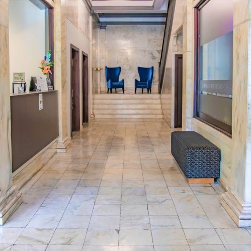 A spacious lobby with marble floors, featuring two blue chairs, a staircase, and a decorative wall.