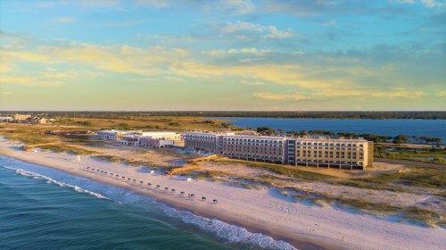 Aerial view of a beachfront hotel with sandy shores and calm waters under a colorful sky.