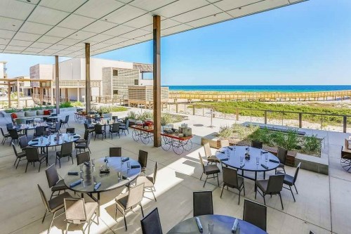 Outdoor dining area with round tables overlooking a beach and ocean under a clear blue sky.
