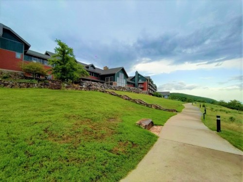 A pathway leads through a grassy area with buildings in the background under a cloudy sky.