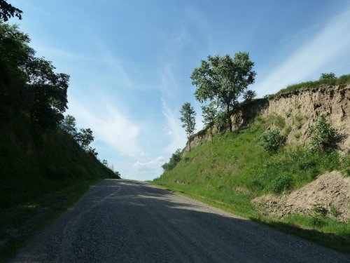 A gravel road flanked by grassy hills and trees under a blue sky with wispy clouds.
