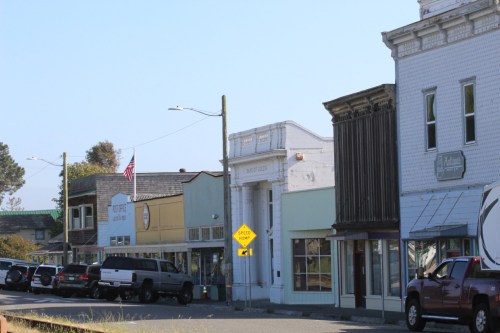 A quaint street lined with small shops and parked cars, featuring a mix of architectural styles and a clear blue sky.