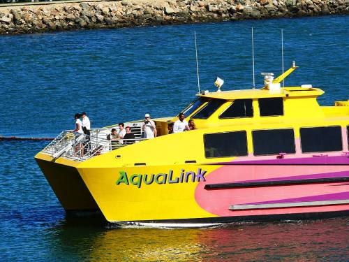 A bright yellow and pink ferry named AquaLink sails on blue water, with passengers enjoying the ride on deck.