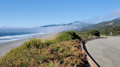 A scenic coastal view with a winding road, sandy beach, and fog rolling over the mountains in the background.
