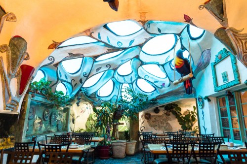 Colorful restaurant interior with unique ceiling design, plants, and tables set for dining. Bright and artistic atmosphere.