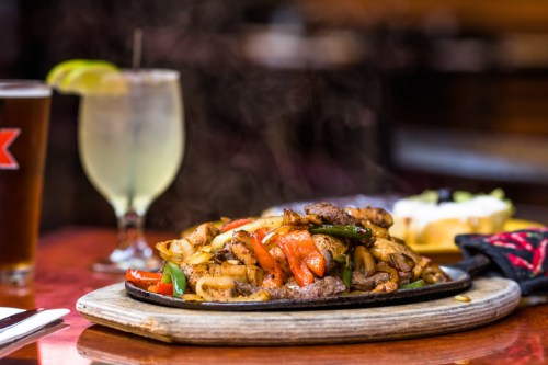 Sizzling fajitas with beef, peppers, and onions on a wooden platter, accompanied by drinks and a side dish.