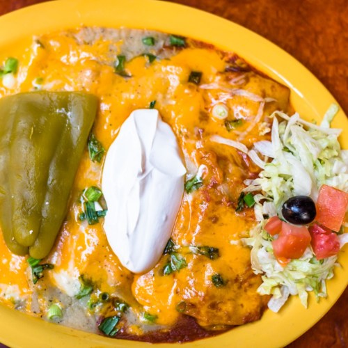 A plate of enchiladas topped with cheese, sour cream, green peppers, and a side of lettuce and tomato salad.