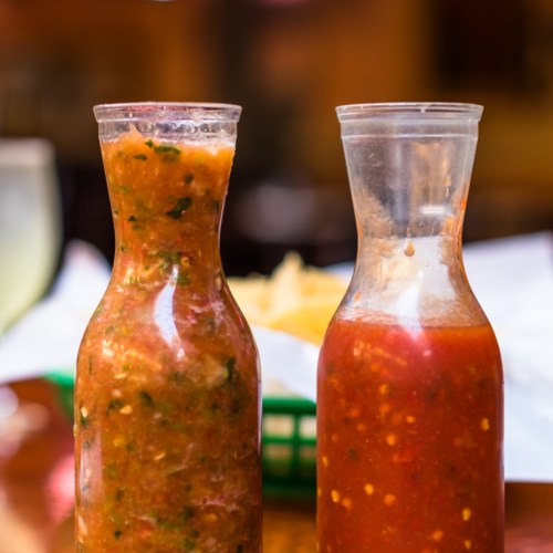 Two glass bottles filled with different types of salsa, one green and one red, on a wooden table.