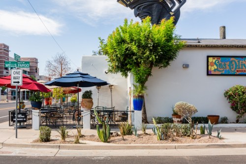 A vibrant restaurant exterior with outdoor seating, colorful umbrellas, and decorative plants under a clear blue sky.