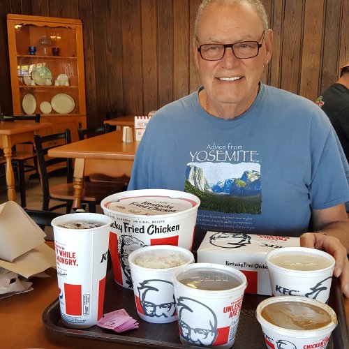A man wearing glasses sits at a table in a restaurant, enjoying a meal from Kentucky Fried Chicken.