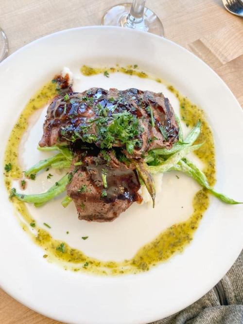 A plate of grilled meat topped with sauce, garnished with herbs, served on a bed of green vegetables.