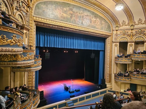 Interior of a grand theater with ornate architecture, featuring a dark stage and audience seating in balconies.