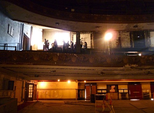 A dimly lit, abandoned theater with people standing on the upper balcony, surrounded by peeling walls and debris.