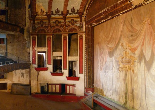 An abandoned theater interior featuring ornate walls, a faded stage curtain, and empty seating areas.