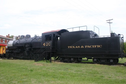 A vintage steam locomotive labeled "Texas & Pacific" parked on a grassy area near a building.