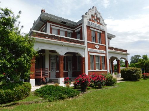 Historic red brick train station with white trim, surrounded by green grass and colorful flower beds.