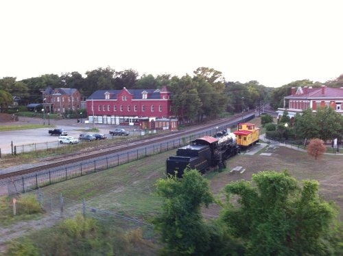 A vintage steam locomotive and yellow caboose sit beside a railway, with red buildings in the background and trees nearby.