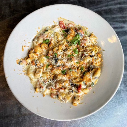 A creamy pasta dish topped with cheese, herbs, and colorful vegetables, served in a white bowl on a metallic surface.