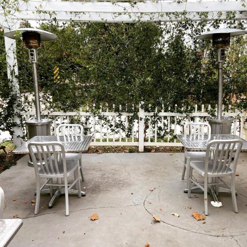Two metal tables and chairs under a covered patio with heaters, surrounded by greenery and a white picket fence.