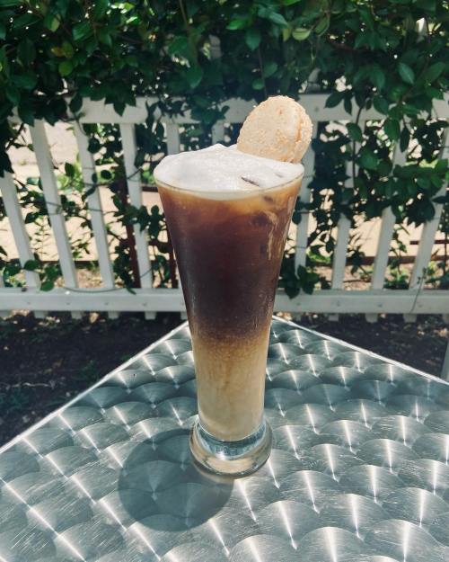A tall glass of iced coffee layered with cream, topped with a cookie, set on a shiny table with greenery in the background.