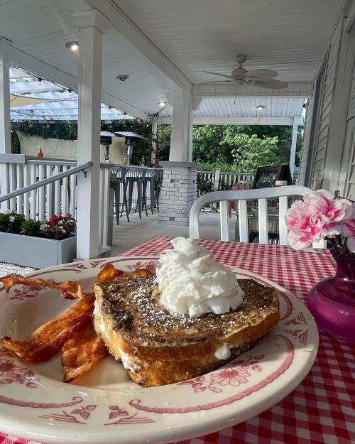 A plate of French toast topped with whipped cream, served with crispy bacon, on a checkered tablecloth in a cozy outdoor setting.
