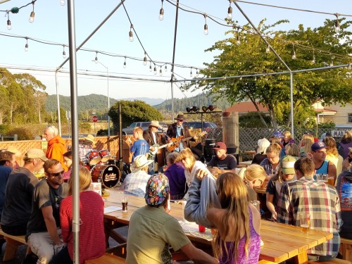 A lively outdoor scene with a band playing on stage, surrounded by a crowd enjoying drinks at picnic tables.