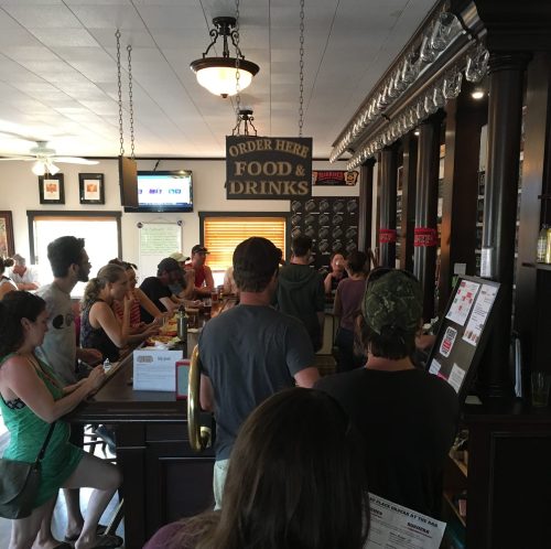 A busy bar scene with patrons waiting to order food and drinks at a counter, lively atmosphere with people chatting.