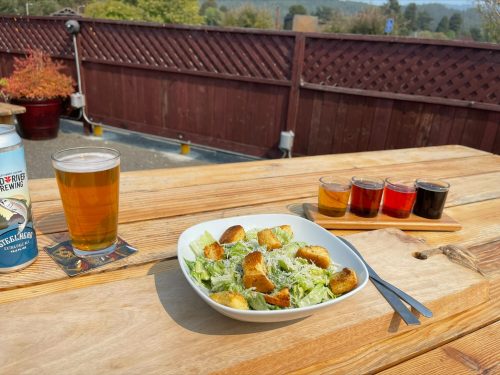 A wooden table with a Caesar salad, a pint of beer, and a flight of four colorful beer samples in the background.