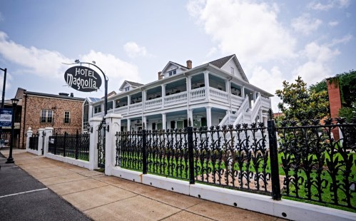 Historic white hotel with a sign reading "Hotel Magnolia," surrounded by a black wrought-iron fence and green grass.