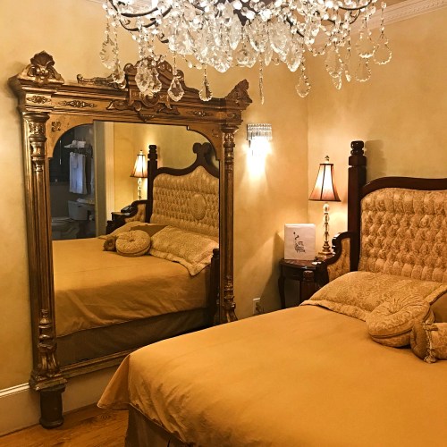 A cozy bedroom featuring a large ornate mirror, a chandelier, and a plush bed with golden bedding and decorative pillows.