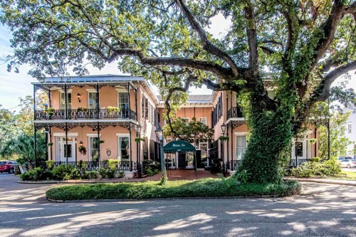 Historic building with balconies, surrounded by lush greenery and a large tree, featuring a welcoming entrance.