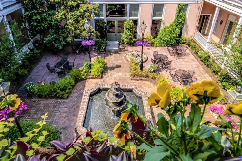 A serene courtyard with a fountain, surrounded by greenery and colorful flowers, featuring outdoor seating areas.